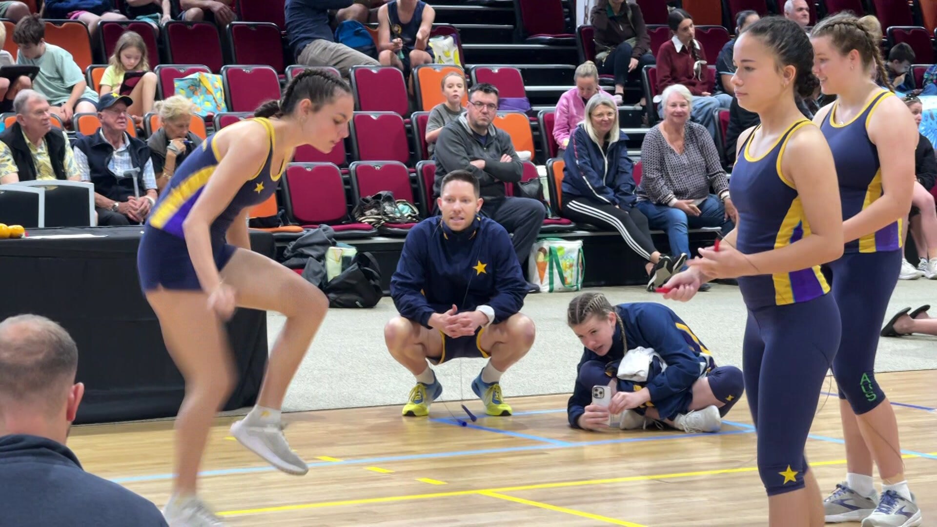 A man crouched down on the sidelines watching women's competitive skipping