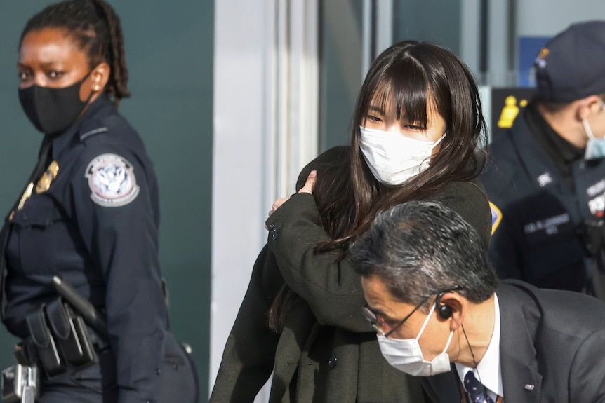A woman wearing a white face mask stands while a female security guard stares on