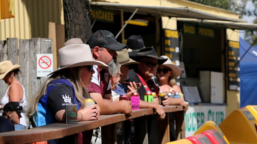 A group of people in cowboy hats rest their drinks on a wooden rail.