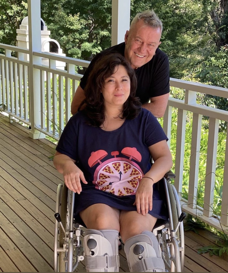 Elly-May sitting in a wheelchair smiling, Jimmy leaning down behind her smiling.