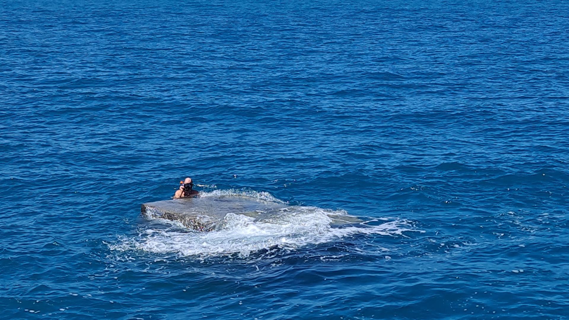 a steel box floating in the sea with a man in a snorkeling mask holding on to it.