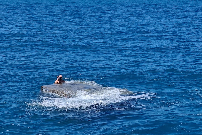 a steel box floating in the sea with a man in a snorkeling mask holding on to it.