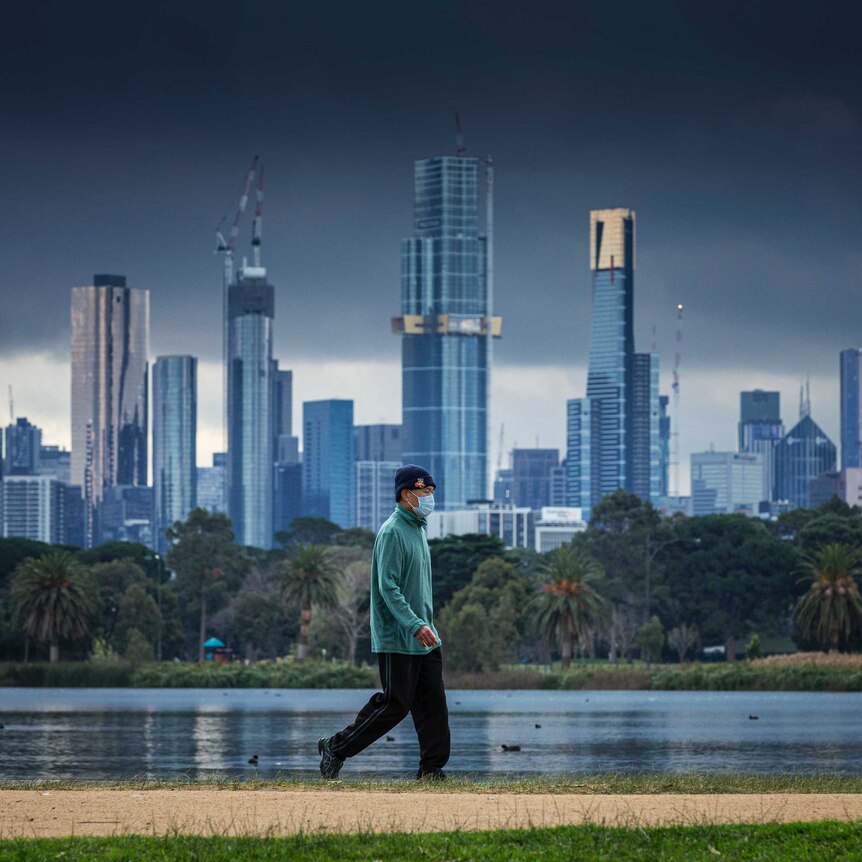 A man wearing a face mask walks by a lake with the city skyline in the background.