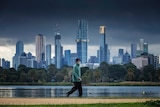 A man wearing a face mask walks by a lake with the city skyline in the background.