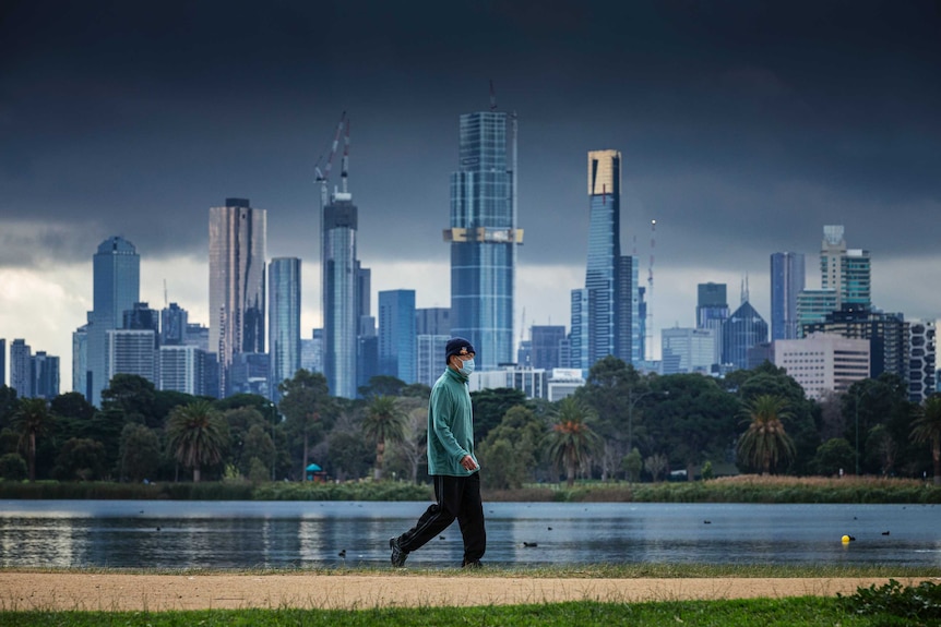 A man wearing a face mask walks by a lake with the city skyline in the background.