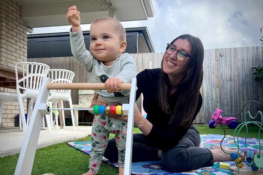 A mum and her baby daughter play with toys on a mat outside