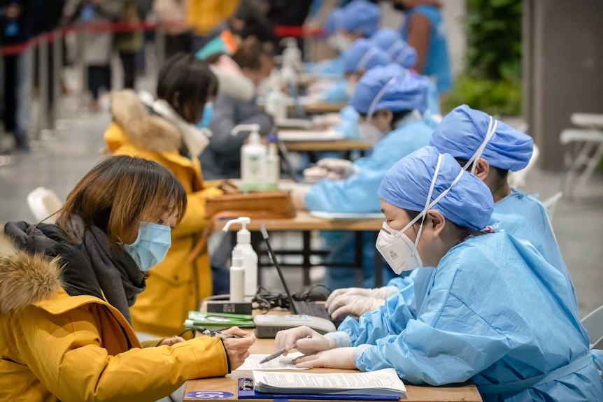 Two people wearing protective clothing and masks faces eachother leaning over a table with documents.