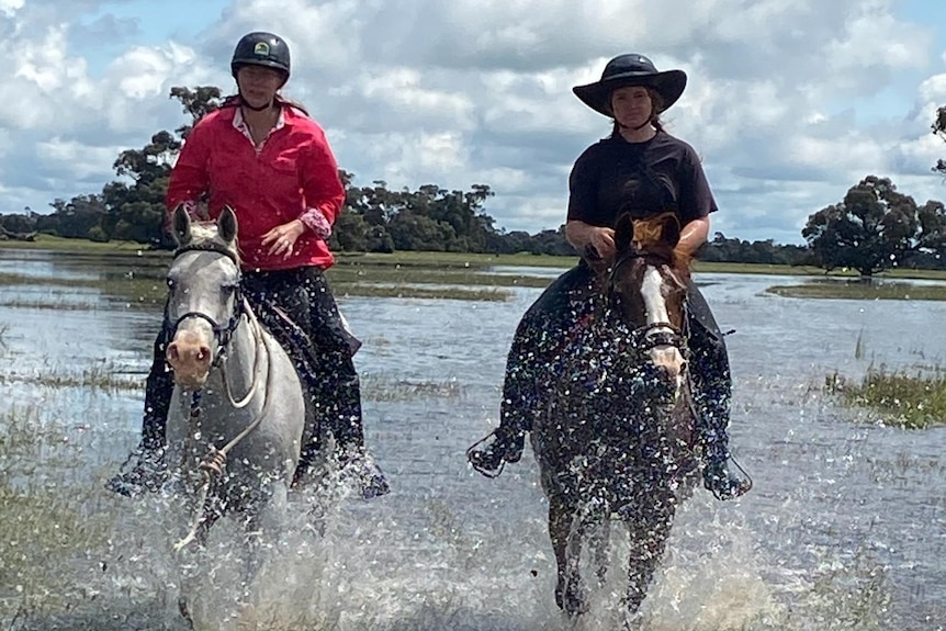 Two people riding horses through floodwater on a farm.