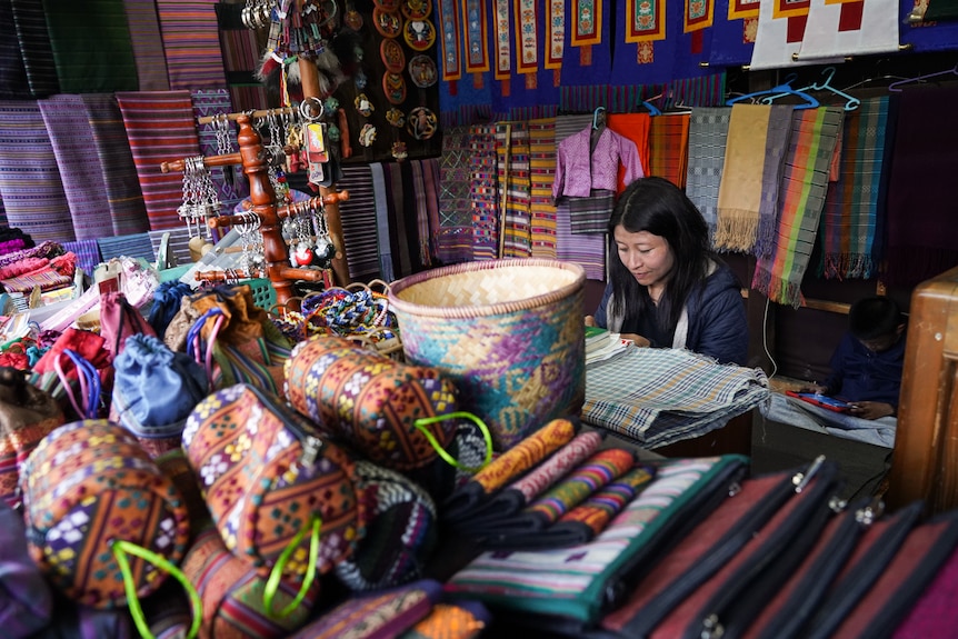 A woman sits weaving a pattern of fabric while surrounded by scarves.