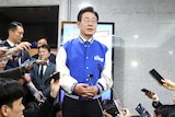 South Korean male politician stands in the centre of a room surrounded by reporters.