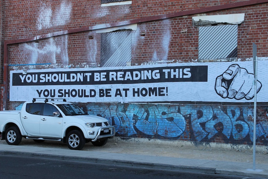 a big sign across a red brick warehouse telling people they should be at home