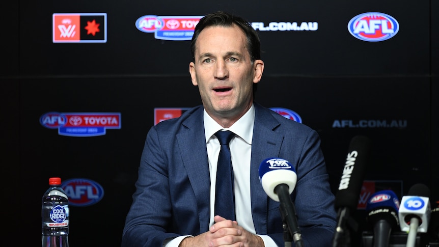 A top AFL executive sits at a desk facing the media with microphones in front of him.