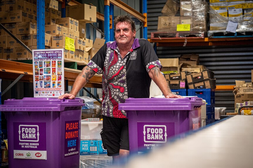 Man in colourful shirt in warehouse holding two purple wheeliebins.
