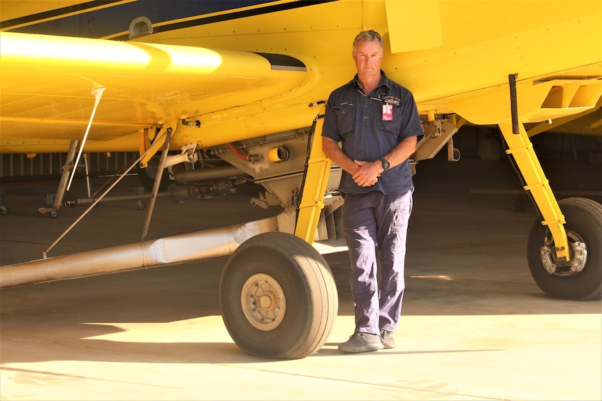 He stands in blue work clothes near a yellow plane, only the wheels and part of the plane's wing are visible 