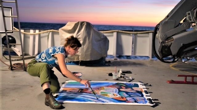 A woman paints on the boat deck surrounded by sea and sunrise.