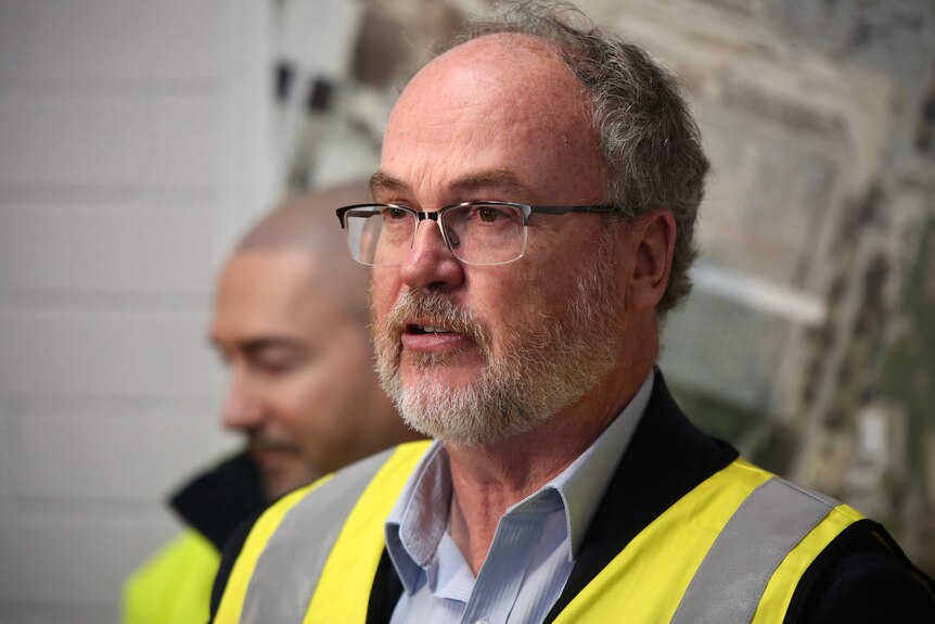 A middle-aged man with a beard wearing a blue button-down and a high visibility vest, looking to left of frame.