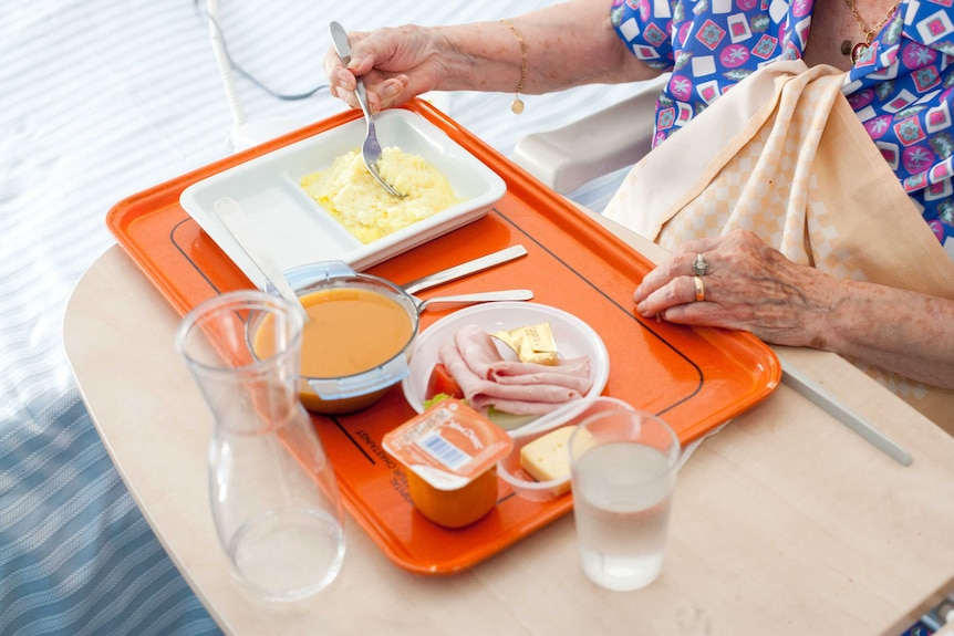 An older woman eats from a tray of bland-looking food, including ham and soup.