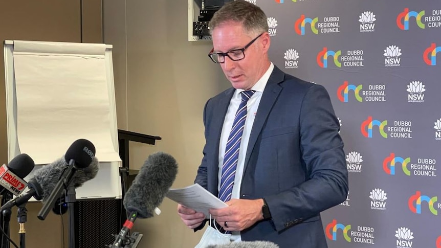 A bespectacled man with short grey hair, wearing a dark suit, addresses the media.