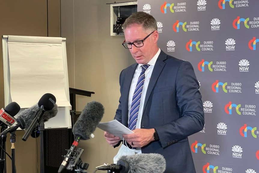 A bespectacled man with short grey hair, wearing a dark suit, addresses the media.