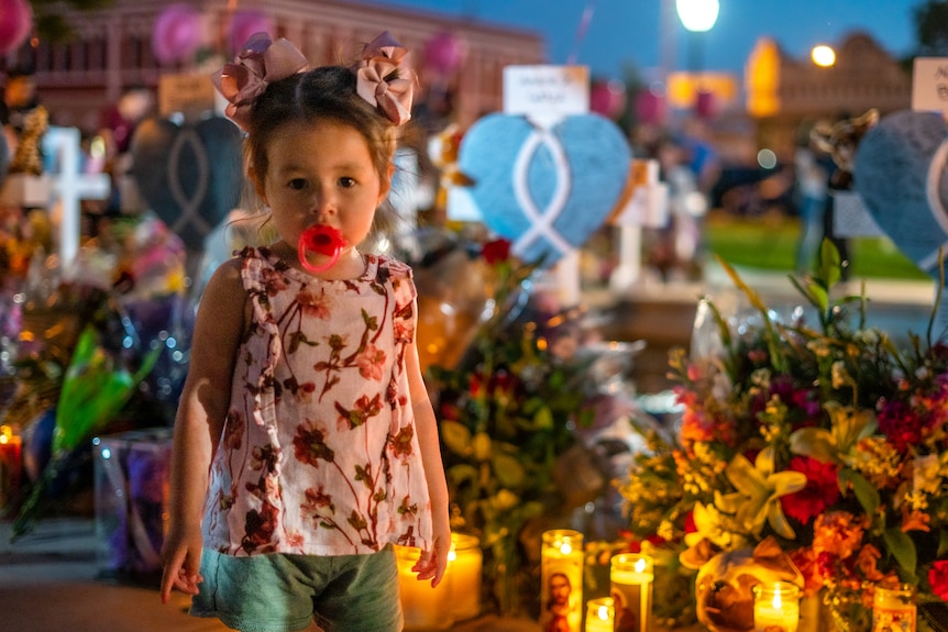 A little girl with a dummy in her mouth stands next to a vigil heaped with flowers and candles 