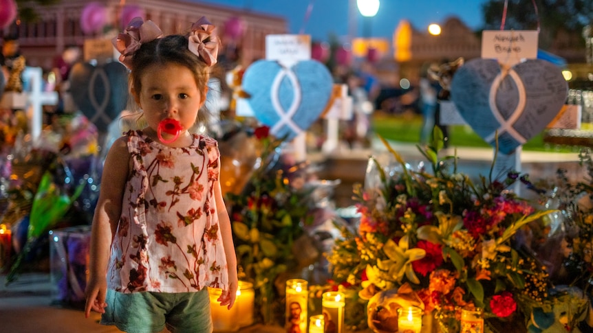 A little girl with a dummy in her mouth stands next to a vigil heaped with flowers and candles 