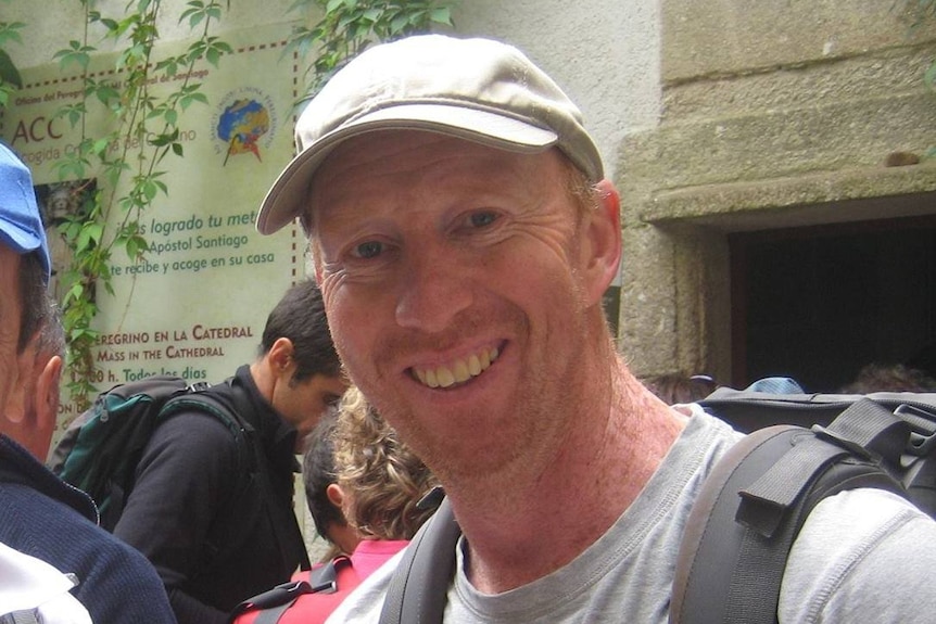 A man wearing a backpack and cap standing outside smiles.