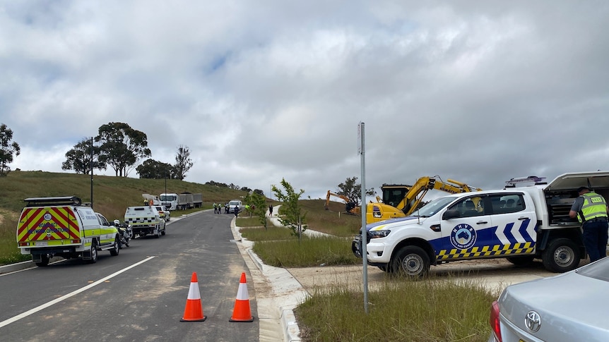 Three emergency services cars line the street and a man looks in his toolkit on the back of his ute