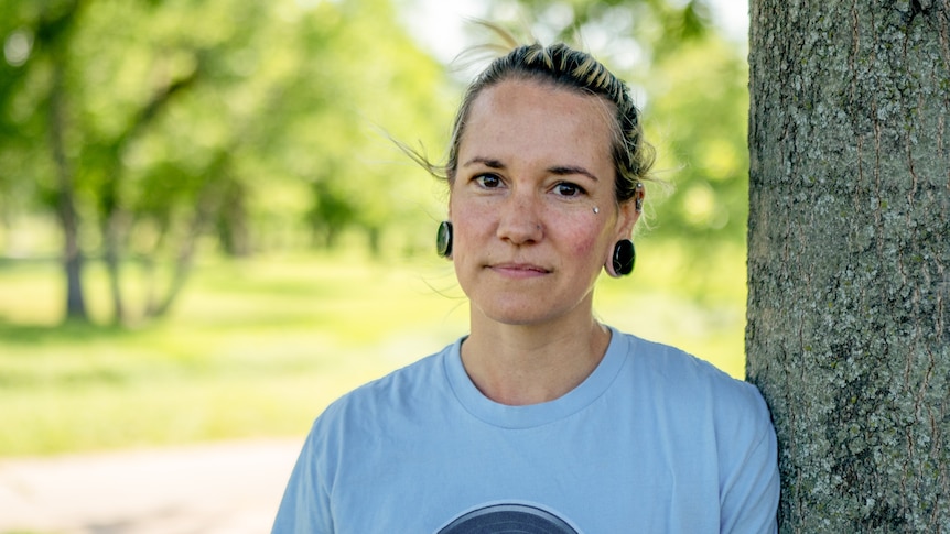 A young woman stands next to a tree with a neutral expression. She wears spacers in her ears and has a dermal piercing