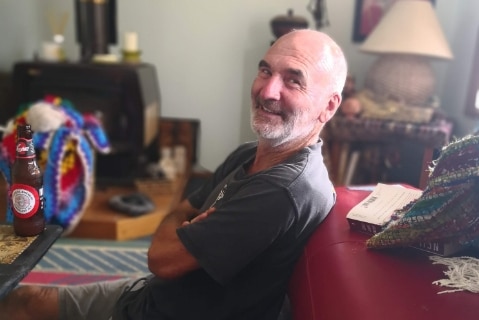 An older man smiles at the camera from a loungeroom.