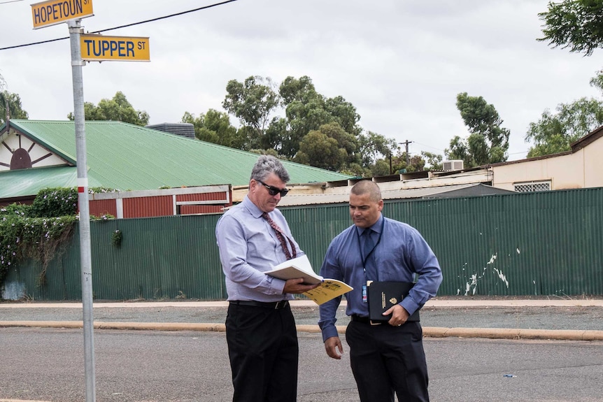 Two detectives stand on a residential street corner in Boulder looking at a notebook.