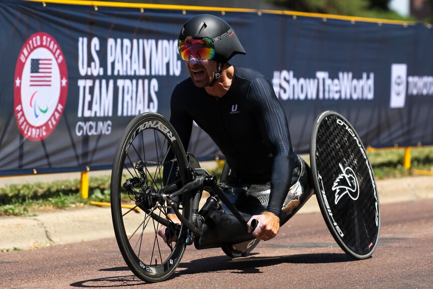 David Berling, wearing a black helmet and cycling gear, races his handcycle past a sign that says 'U.S. Paralympic Team Trials'