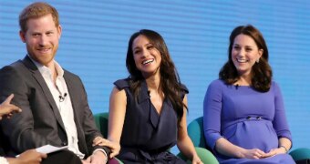 Prince Harry wears a black suit as he sits on chair next to Meghan Markle in  black dress and Kate Middleton in blue.