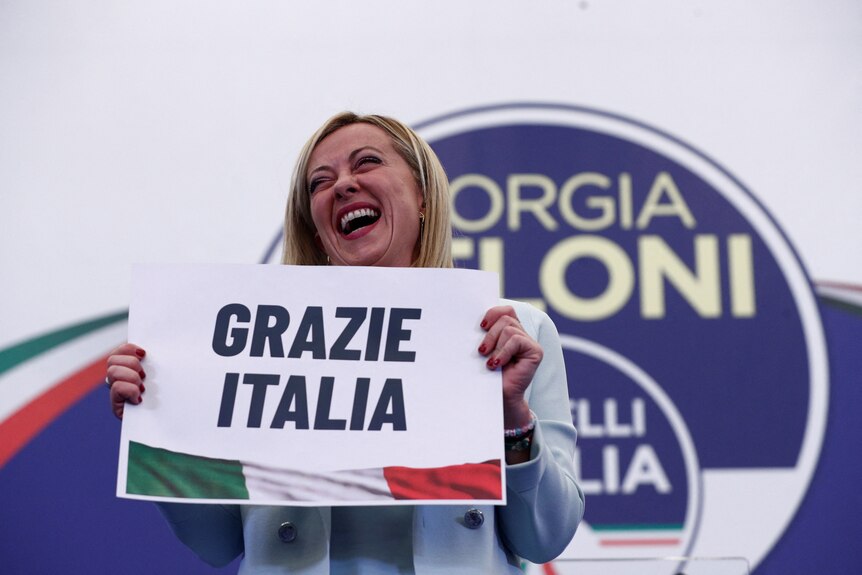 A blonde middle-aged woman smiles and holds a poster reading 'grazie Italia'