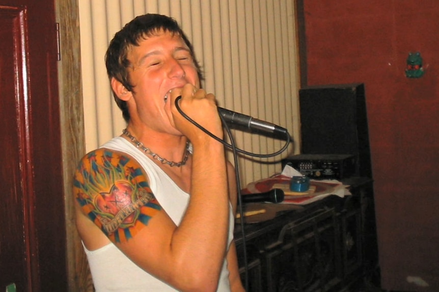 A teenager with a large tattoo on his forearm sings into a microphone, his eyes closed 