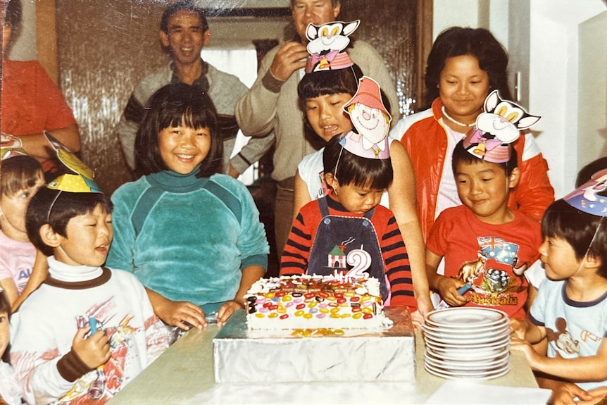 A film photo of a group of kids at a birthday party with a child in the middle facing a birthday cake.