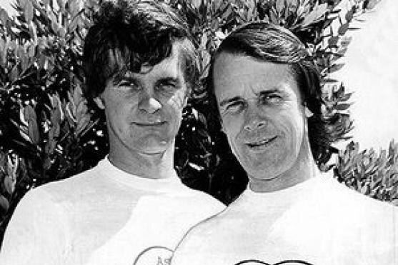 A black and white photo of two slightly smiling young men, both wearing same white tee-shirt, stand in front of trees.