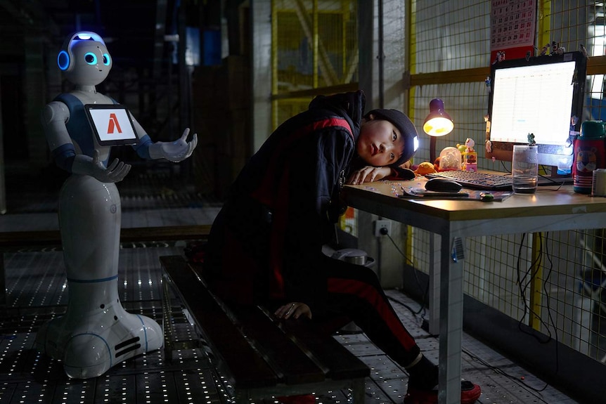 Film still featuring young person slumped over desk in front of computer, with robot standing behind, arms outstretched.