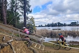 Two cyclists in lycra carry their bikes up a set of stairs beside a lake with reeds and blue sky.