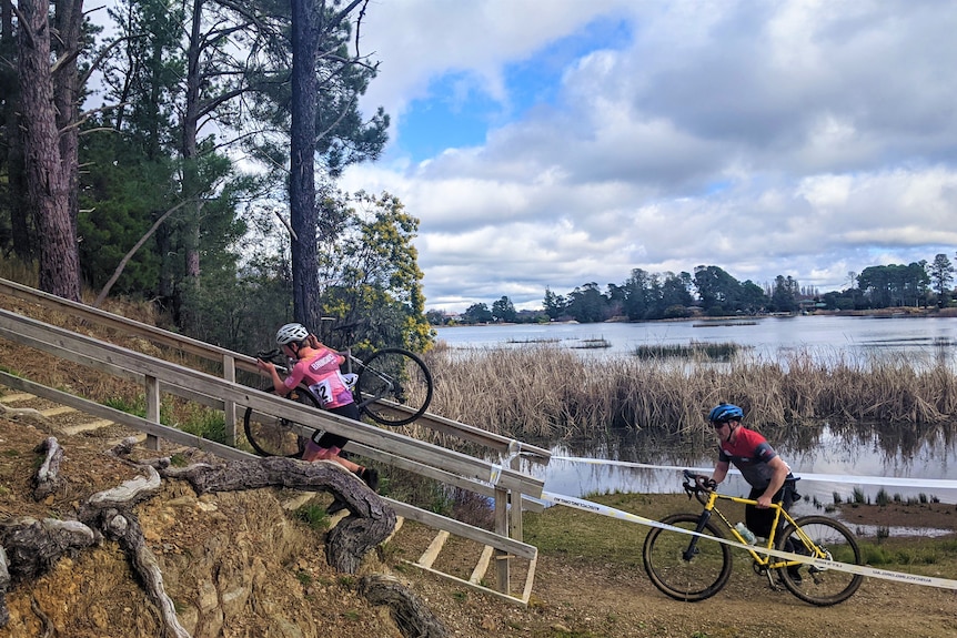 Two cyclists in lycra carry their bikes up a set of stairs beside a lake with reeds and blue sky