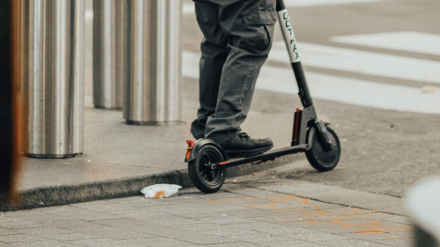 A man wearing grey cargo pants and a grey jacket stopped on a e-scooter on the corner of a city street.
