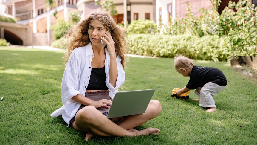 A young woman working outside on a laptop and phone, while a toddler plays next to her. 