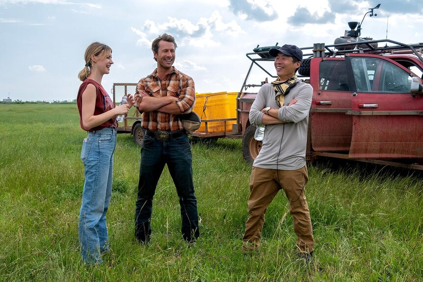 Three people stand on a film set on green farmland next to an SUV in a behind-the-scenes film shot.