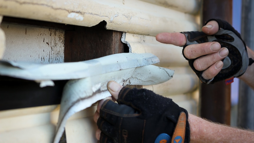 Roden Woodhams peels back tape to reveal a gaping hole in the wall through which asbestos can be seen.