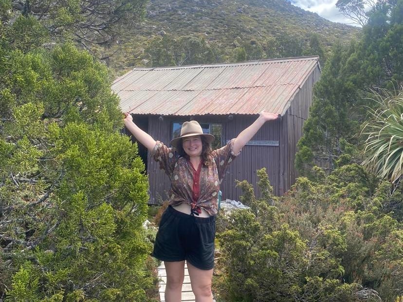 Person with mountain hut behind them and wide arms smiles at camera