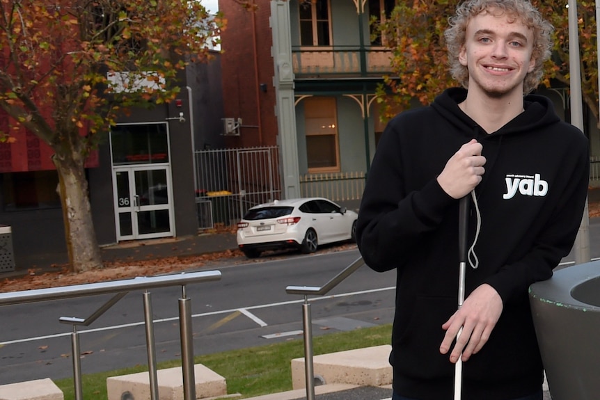 A young man who is vision impaired wears a black jumper that says 'YAB' in a Ballarat street.