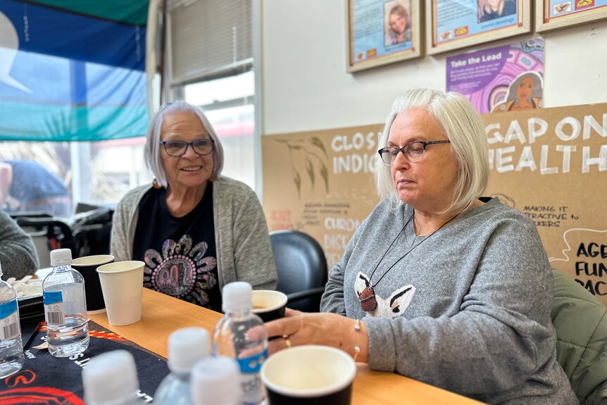 Two ladies share cups of tea at an Aboriginal health service. They each have grey hair, bob haircuts.
