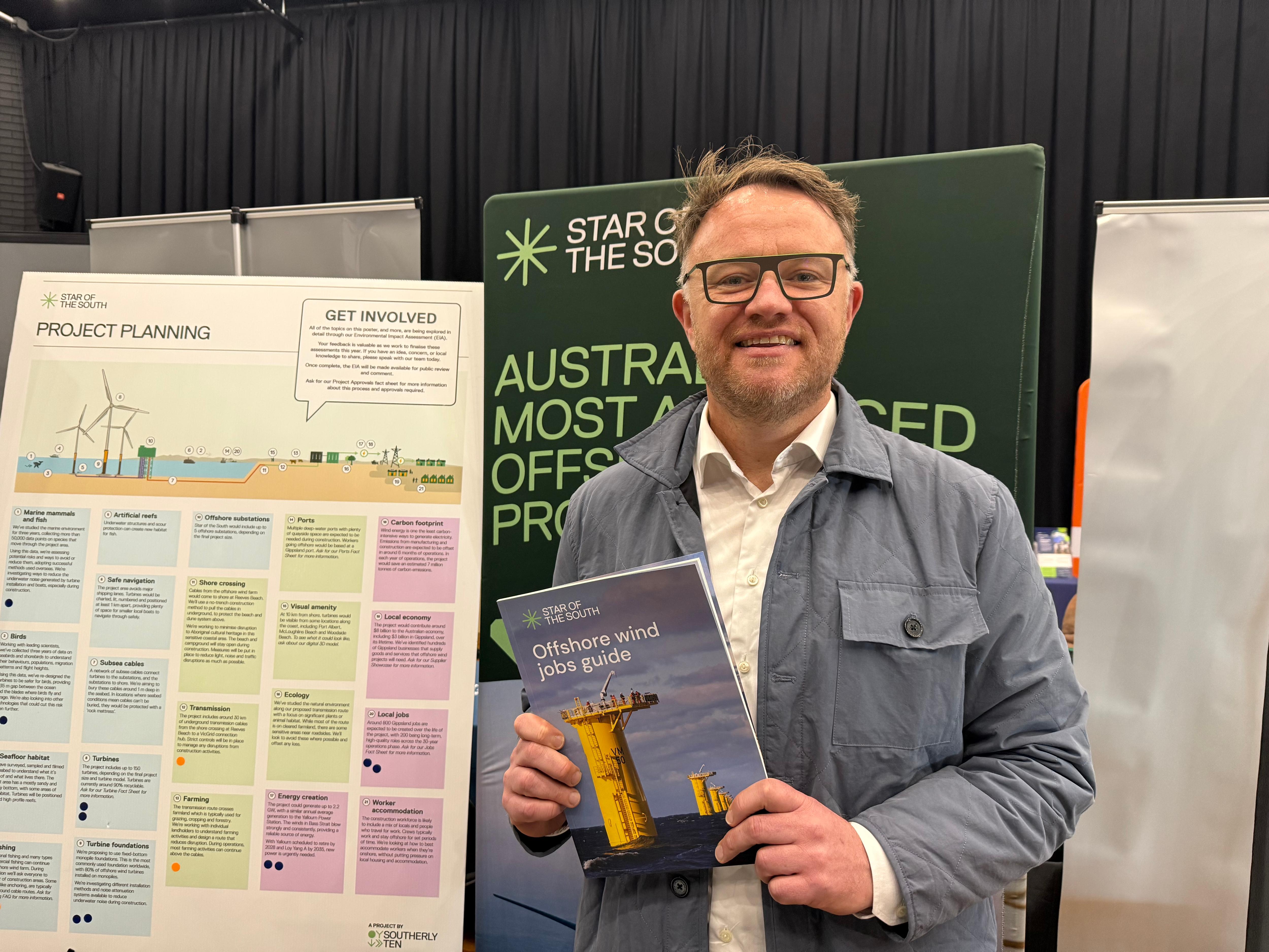 A man wears a grey suit and glasses, standing in front of an offshore wind information stand. 