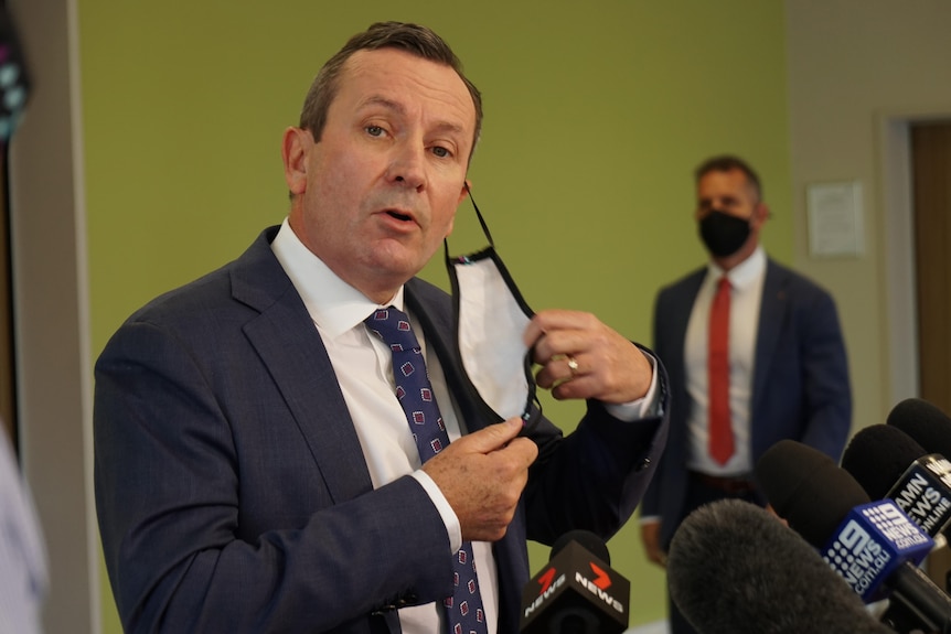 A mid shot of WA Premier Mark McGowan removing a face mask for a media conference indoors.