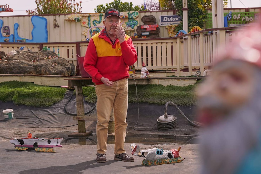 A man in a red and yellow jumper stands in a large empty pond with old, tattered toy boats lying on the ground around him.