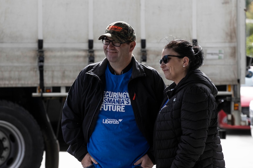 A man in a blue shirt wearing glasses, a cap and a jacket stands next to a woman